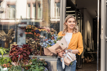 Wall Mural - attractive smiling girl going out from flower shop with wrapped bouquet and looking away