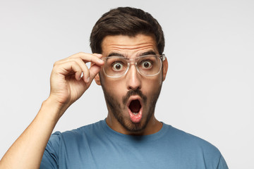 Studio portrait of young european caucasian man isolated on gray background, dressed in blue t-shirt, holding transparent plastic glasses looking with round open mouth with surprise