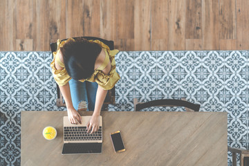 Top view of young asian woman sitting, using laptop in modern workplace, café coffee shop. Urban female college lifestyle concept on wooden table, blue vintage floor tile pattern with copy space.