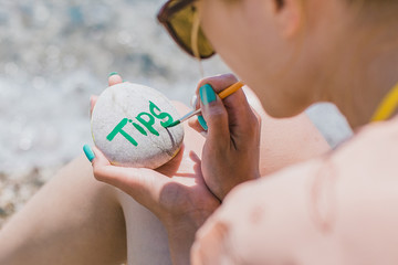 Girl painting with brush on pebble stone  sign tips. Tips. Sea holidays. Sea pebbles on the beach background