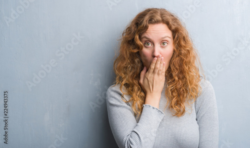 Fotomural Young redhead woman over grey grunge wall cover mouth with hand shocked with sha