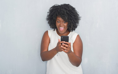 Canvas Print - Young african american woman over grey grunge wall texting a message using smartphone scared in shock with a surprise face, afraid and excited with fear expression