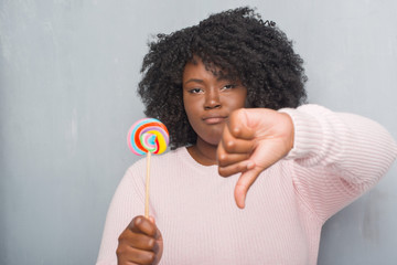 Sticker - Young african american woman over grey grunge wall eating lollipop candy with angry face, negative sign showing dislike with thumbs down, rejection concept