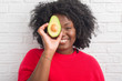 © Krakenimages.com - Young african american woman over white brick wall eating avocado with a happy face standing and smiling with a confident smile showing teeth