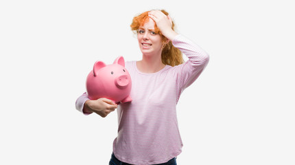 Poster - Young redhead woman holding piggy bank stressed with hand on head, shocked with shame and surprise face, angry and frustrated. Fear and upset for mistake.