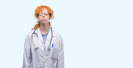 Wall Mural - Young redhead woman wearing doctor uniform puffing cheeks with funny face. Mouth inflated with air, crazy expression.