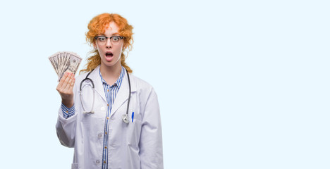 Poster - Young redhead doctor woman holding dollars scared in shock with a surprise face, afraid and excited with fear expression