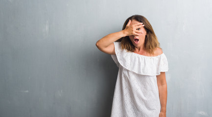 Poster - Middle age hispanic woman standing over grey grunge wall peeking in shock covering face and eyes with hand, looking through fingers with embarrassed expression.