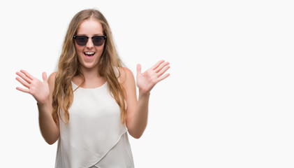Wall Mural - Young blonde woman wearing sunglasses celebrating crazy and amazed for success with arms raised and open eyes screaming excited. Winner concept
