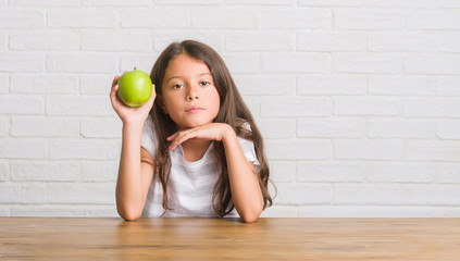 Poster - Young hispanic kid sitting on the table eating fresh green apple with a confident expression on smart face thinking serious