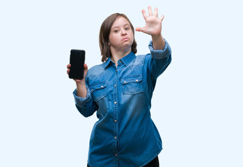 Wall Mural - Young adult woman with down syndrome showing smartphone screen over isolated background with open hand doing stop sign with serious and confident expression, defense gesture