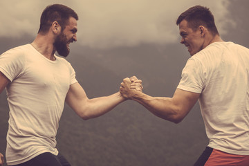 Strong male bodybuilders in white t-shirts greeting each other in wrestling manner outdoor over foggy mountain landscape. Rivalry, challenge, strength comparison concept