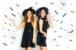 © maxbelchenko - portrait of two happy young women in black witch halloween costumes on party over white background with bats . firecrackers in the background. confetti . the concept of Halloween . funny faces.