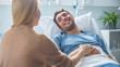 © Gorodenkoff - In the Hospital Sick Man Lying on the Bed, His Visiting Wife is Sitting Beside Him and Talking, Holding His Hands.