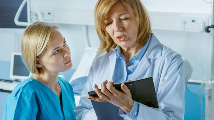 Wall Mural - Professional Female Doctor and Young Nurse Discuss Patient's Chart. In the Background Sick Man Lying in the Bed Waiting to be Cured.