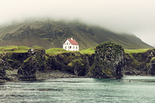 Lonely Icelandic House With Red Roof On The Sea Coast With Green Grass Meadow, Rocks Anf Foggy Sky. Natural Iceland Travel Landscape.