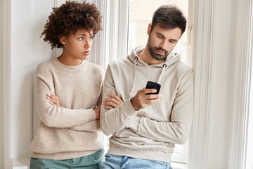 Photo of mixed race lovers dressed in casual sweatshirts, stand together near window, sort out relationships. Jealous ethnic woman keeps arms folded, looks with sad expression at guy who uses gadget