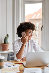 Vertical shot of cheerful woman bookkeeper talks on mobile phone, has positive expression, dressed in white stylish blouse, works with laptop computer, has work break, drinks hot aromatic beverage