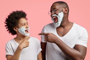 Wall Mural - Horizontal shot of happy son and dad have fun together, indicate at each other, laugh joyfully, have shaving foam on cheeks, foolish indoor, have morning routine, isolated over pink background