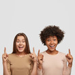 © wayhome.studio  - Joyful beautiful mixed race women have satisfied expressions, point upwards, suggest use copy space wisely, dressed casually, pose over white studio wall. Black girl stands near Caucasian friend