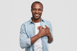 © WHstudio Leushin N - Touched and thankful African man holds hands on chest, expresses gratitude, looks pleasantly at camera, has positive smile, dressed in stylish clothing, poses over white background. Body language
