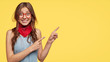 Horizontal shot of satisfied positive woman seller advertises new trends, wears fashionable bandana and denim dress, poses over yellow blank wall with free space for your advertising content.