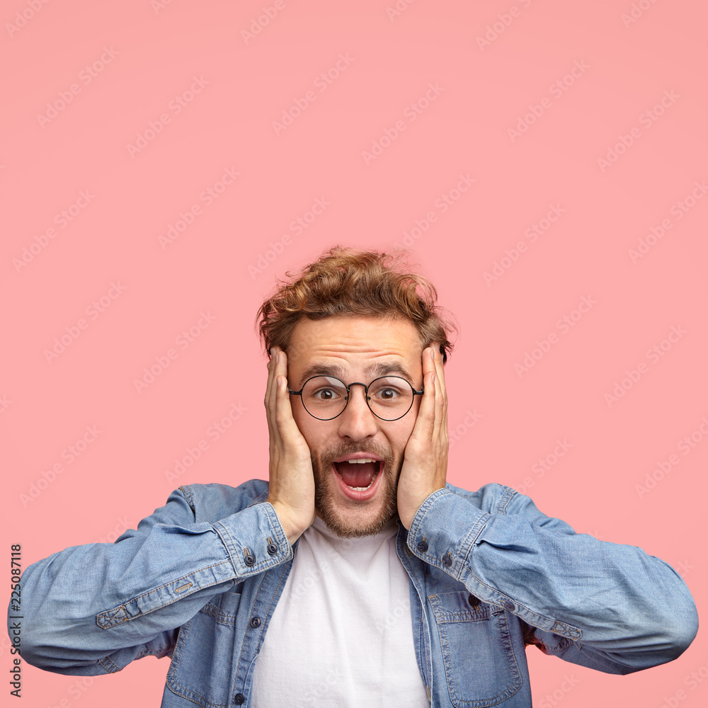Amazed happy guy with overjoyed expression, keeps hands on face, gazes at camera with unexpected cheerful look, has curly hair, dressed in fashionable clothes, poses over pink background, free space - obrazy, fototapety, plakaty 