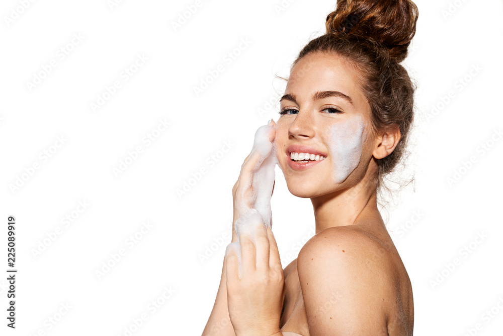 Portrait of young pretty girl with healthy skin on white background. Woman smiling and applying cosmetic foam on face.  Beauty skincare concept. - obrazy, fototapety, plakaty 