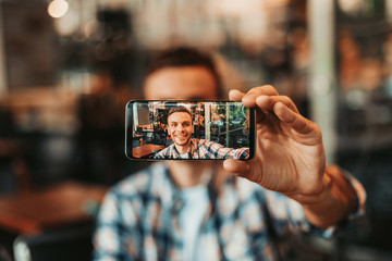 Portrait of positive man taking selfie on mobile while looking at it. Positive guy keeping gadget in hand during fun concept