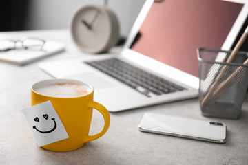 Sticky note with funny face attached to cup of coffee on office table. Space for text