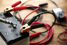 A Car Battery With Red And Black Battery Jumper Cables With Copper Clamps Attached To The Terminals.