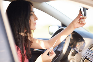 Brunette lady paints lips with lipstick, looks in mirror of car, waits on traffic light, poses in own vehicle. Beautiful businesswoman prepares for date in automobile. Beauty and lifestyle concept