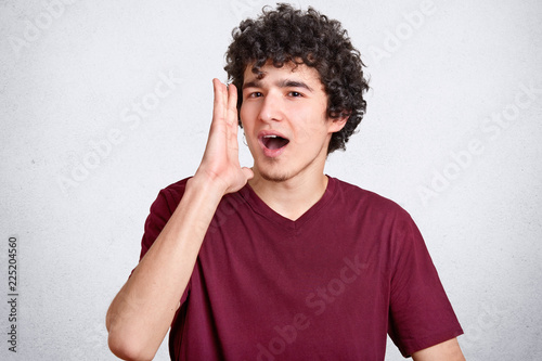 Fotografie, Obraz Portrait of attractive curly European guy keeps hands near widely opened mouth, shouts something, wants somebody hear him, dressed casually, poses over white concrete wall
