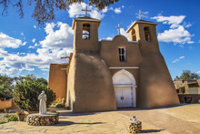 Historic Adobe San Francisco De Asis Mission Church In Taos New Mexico In Dramatic Late Afternoon Light Under Intense Blue Sky With Fluffy While Clouds And Birds In The Belfry