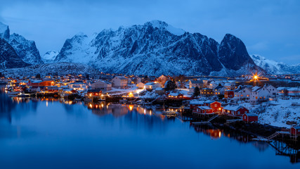 Wall Mural - Lofoten islands, Norway. Colorful winter landscape in blue hours. Illuminated fishing village reflected in water. Snowy mountains in background. 