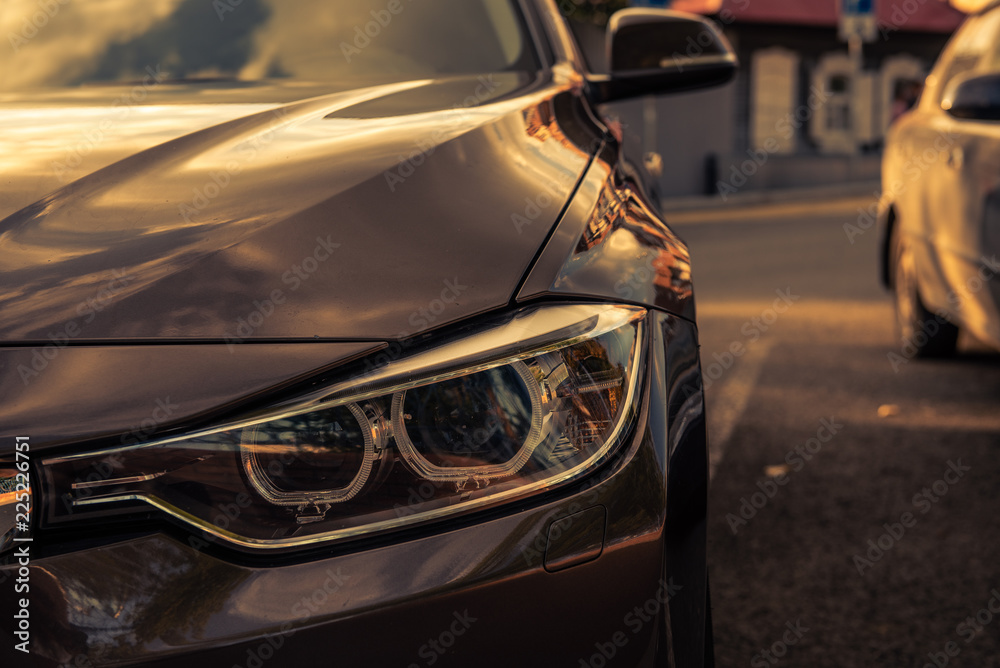 grille and headlight of a brown car in a parking lot against the background of a blurred old house with morning sunbeams - obrazy, fototapety, plakaty 