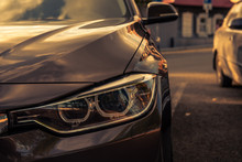 Grille And Headlight Of A Brown Car In A Parking Lot Against The Background Of A Blurred Old House With Morning Sunbeams
