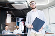© Seventyfour - Content confident young bearded speaker of business training holding clipboard with thesis and looking at camera in conference room
