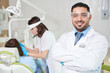 © Seventyfour - Waist up portrait of confident Middle-Eastern doctor wearing lab coat posing in dentists office with patient in background, copy space