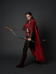 full length portrait of brunette girl wearing red medieval costume and cloak, holding a bow and arrow. standing pose on grey studio background.