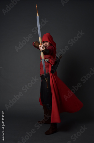 Photographie full length portrait of brunette girl wearing red medieval costume and cloak, holding a bow and arrow