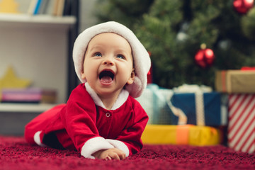 close-up portrait of laughing little baby in santa suit lying on red carpet in front of christmas tr