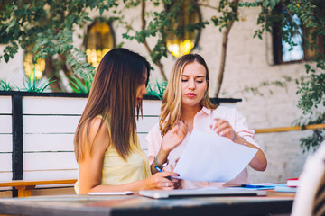 Two businesswomen reading paper documents and using laptop computer in cafe. Economist showing data infographic to colleague analyzing income of company while sitting in cafe