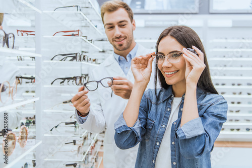 Fotomural happy woman choosing eyeglasses while male oculist standing near with another ey