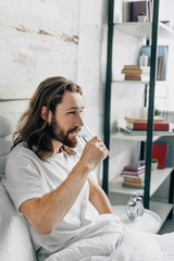 side view of young bearded man with long hair having headache and taking pill with glass of water in bedroom at home