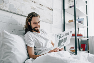 Wall Mural - smiling bearded businessman with long hair reading newspaper in bed during morning time at home