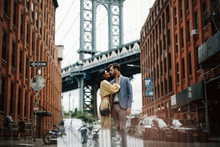 Love Story In New York. Gorgeous Couple Of American Man With Beard And Tender Eastern Woman Hug Each Other Before The Cityscape Of Brooklyn Bridge Somewhere In New York