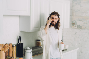 Wall Mural - smiling Jesus in crown of thorns standing with cup of coffee and talking on smartphone in kitchen at home