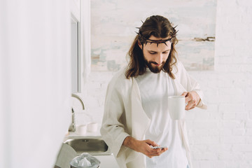 Wall Mural - happy Jesus in crown of thorns standing with cup of coffee and using smartphone in kitchen at home