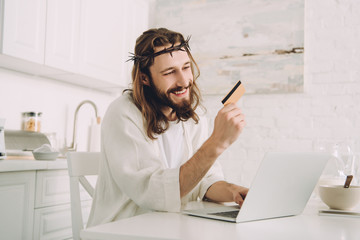 Wall Mural - smiling Jesus holding credit card and doing online shopping at table with laptop in kitchen at home